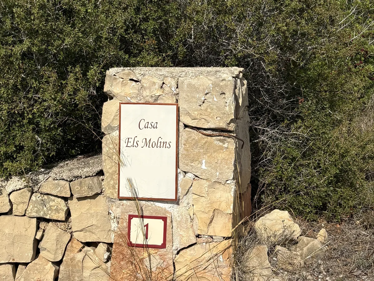 Cartel de piedra y cerámica con el nombre Casa Els Molins junto al camino de acceso